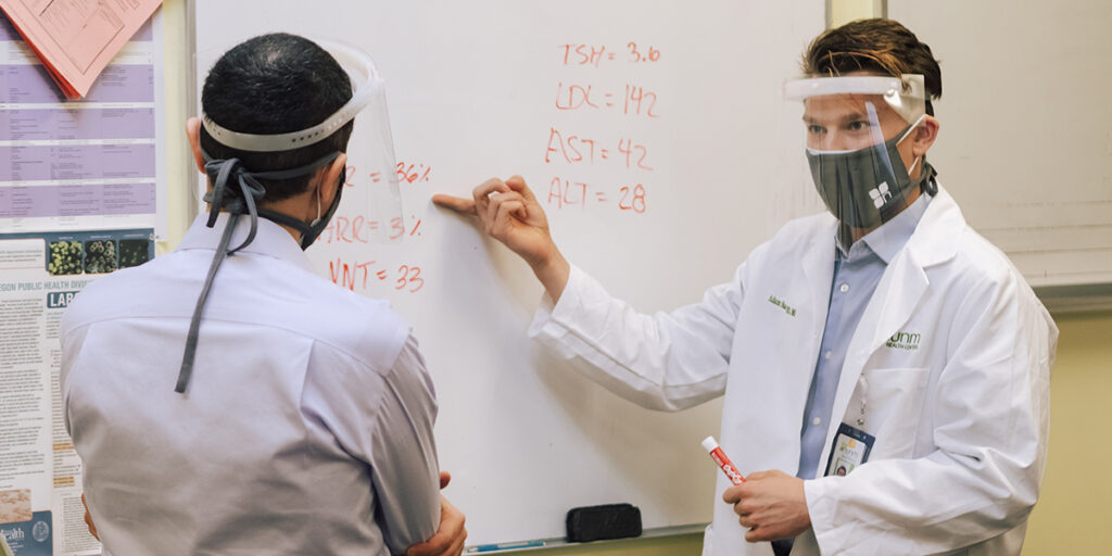 An NUNM student and professor wearing face coverings and eye shields discuss data sets during class on campus. Face coverings are required in all indoor and outdoor settings on NUNM's campus in Portland, Oregon.