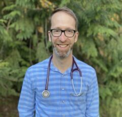 Dr Christopher Randolph smiling for a photo wearing a stethoscope and blue striped shirt with trees in the background of the photo