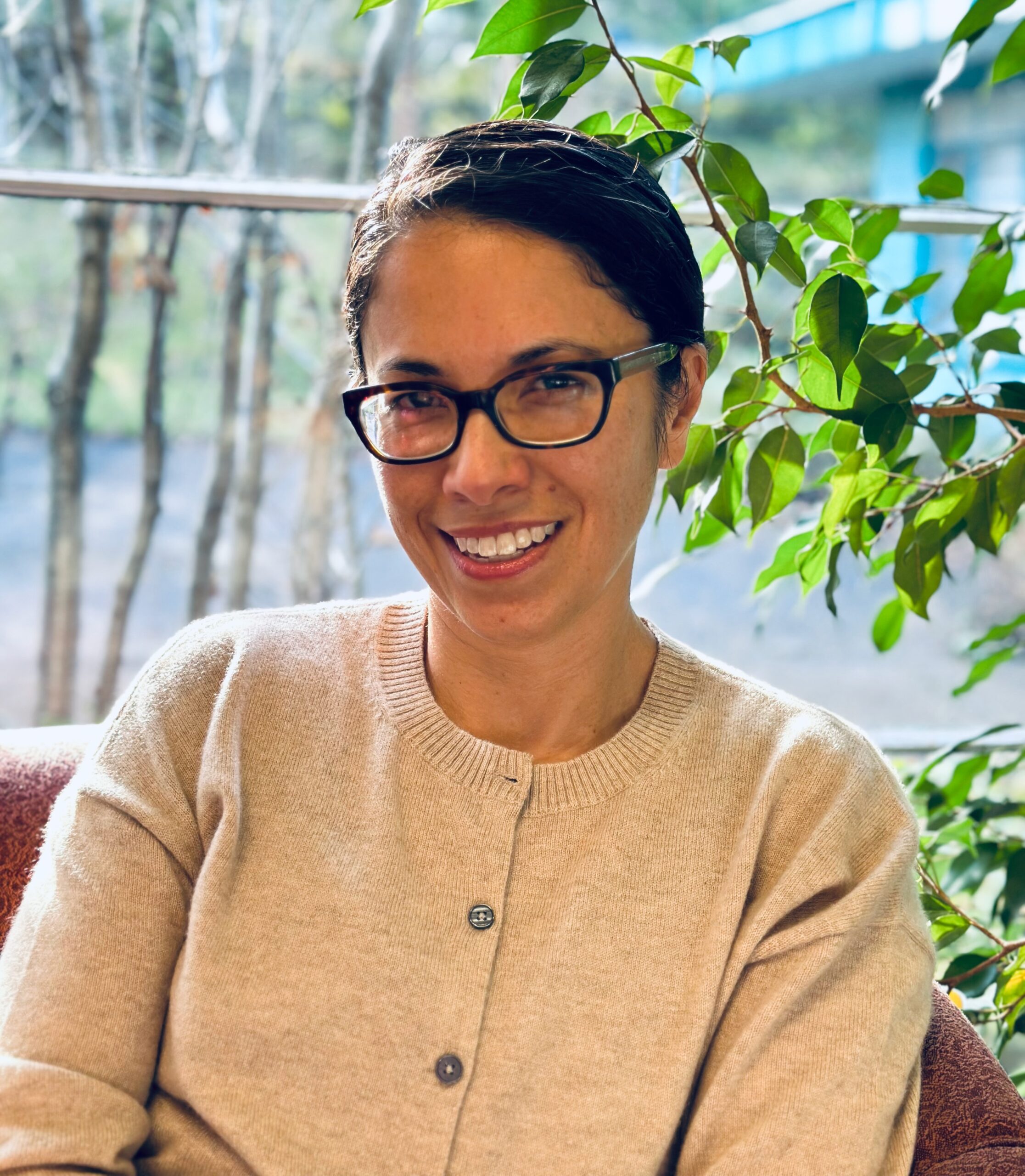Claudia Nakama smiling wearing glasses and a white sweater with leaves and branches from a plant in the background