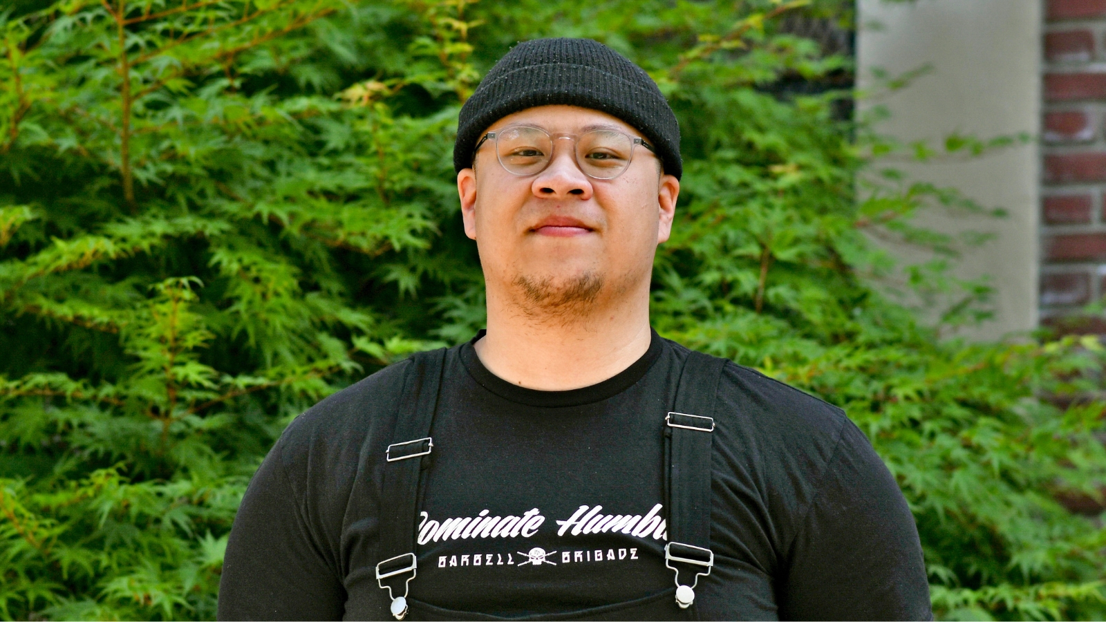 Johnny Lemau, NUNM alumni smiling for a photo wearing a black shirt and black hat with trees in the background