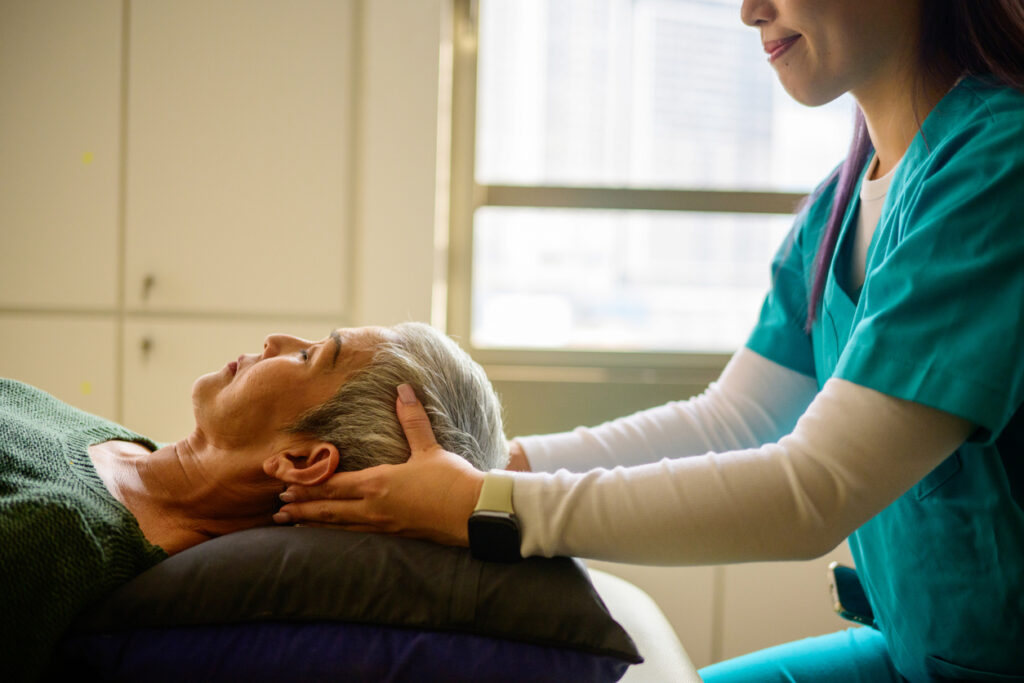 A woman in scrubs, training to be a licensed massage therapist, massages a patient's head as they lie, eyes closed on the table.