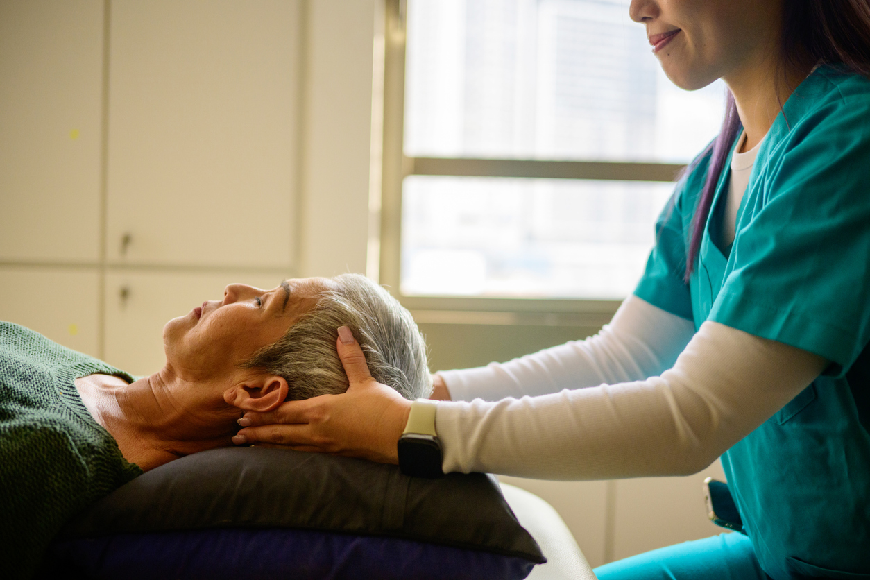 A woman in scrubs, training to be a licensed massage therapist, massages a patient's head as they lie, eyes closed on the table.