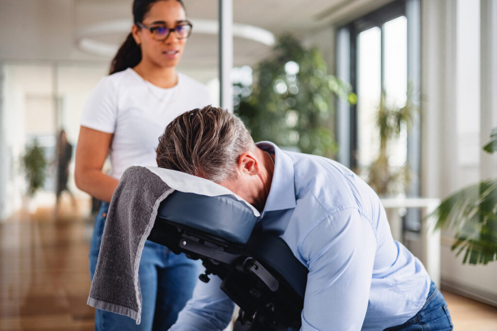 A student, standing over a patient in a massage chair, trains to be a massage therapist.