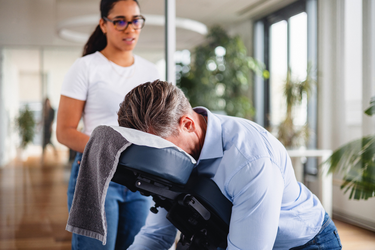 A student, standing over a patient in a massage chair, trains to be a massage therapist.